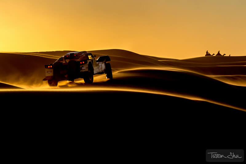 car racing through the Merzuga Desert with camels in the distance